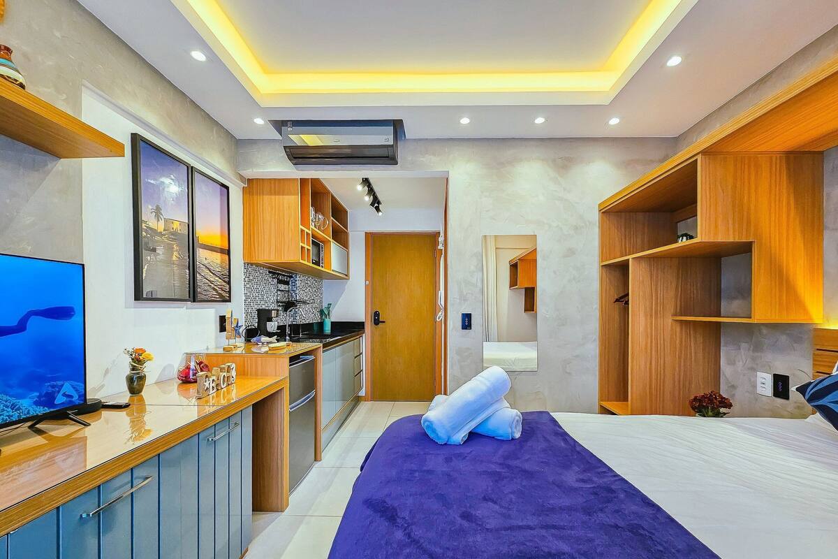 Habitació d'hotel moderna amb petita cuina, un llit amb manta morada i un televisor al costat esquerre.