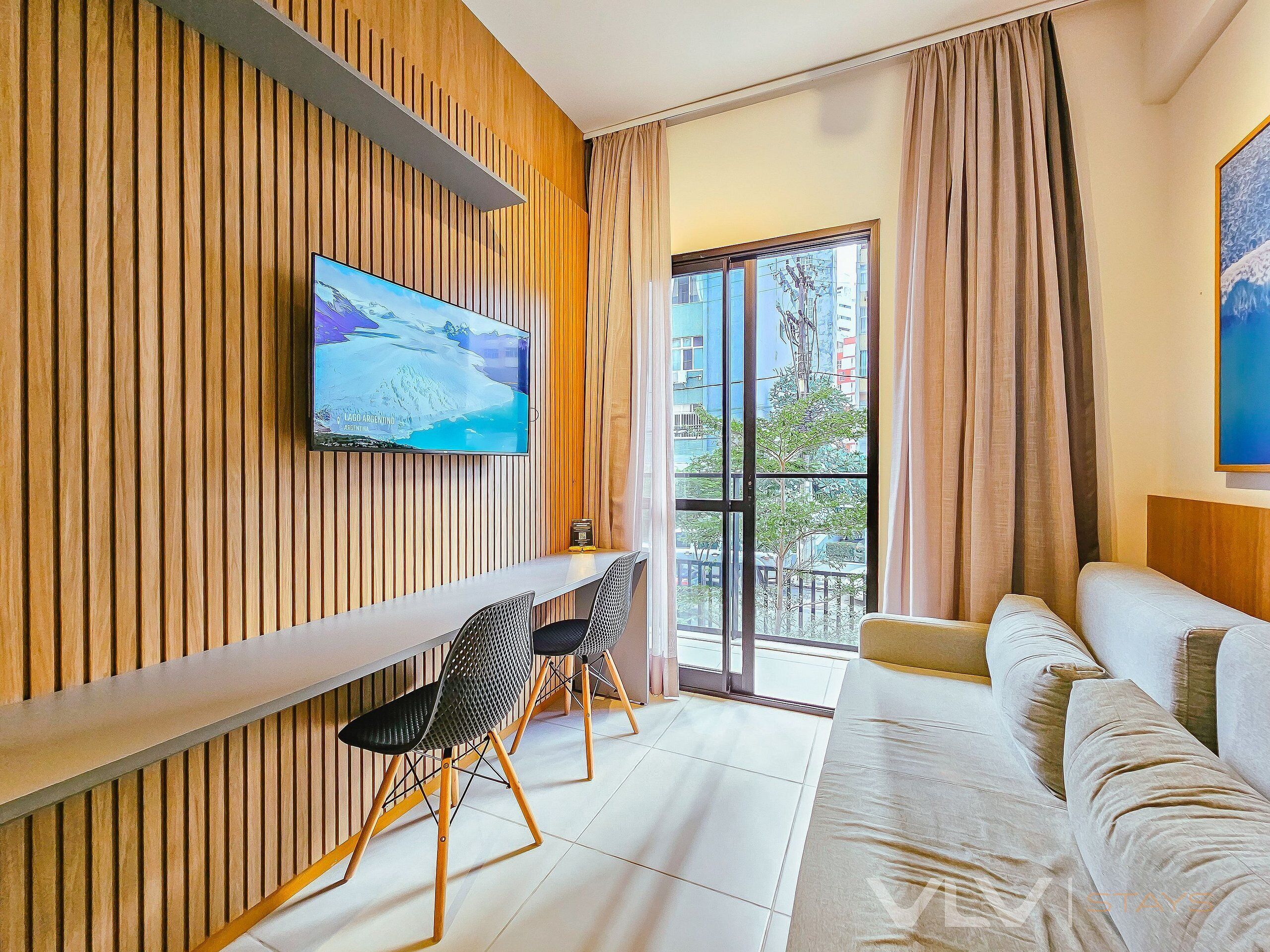 Une chambre d’hôtel moderne avec un mur lambrissé en bois, un téléviseur à écran plat, un canapé, une table à manger avec des chaises et une grande fenêtre avec des rideaux.