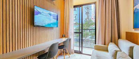 Une chambre d’hôtel moderne avec un mur lambrissé en bois, un téléviseur à écran plat, un canapé, une table à manger avec des chaises et une grande fenêtre avec des rideaux.