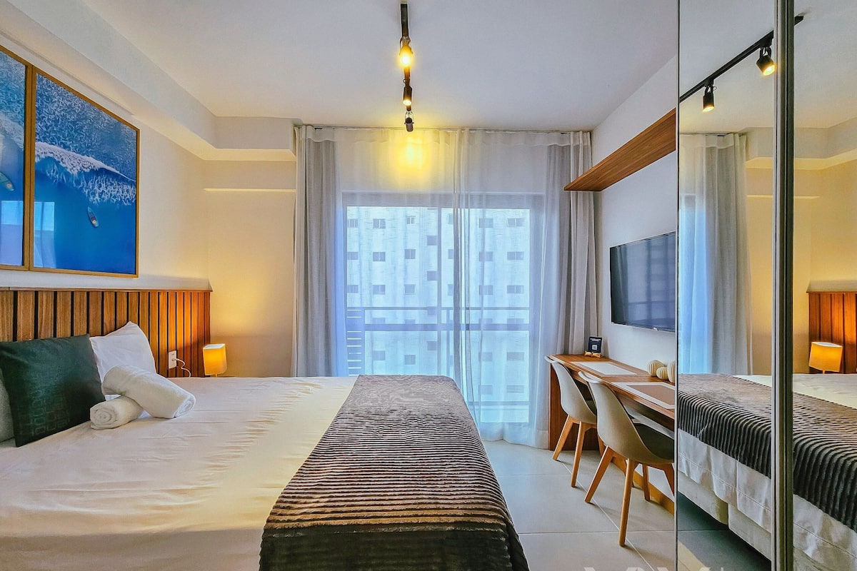 Habitació d'hotel amb llit, escriptori, cadira, televisió i una finestra gran amb cortines.