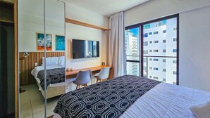 A hotel room with a bed, a desk, a chair, a TV, and a large window with a city view. - Porto Privilege by VLV Stays (Salvador)