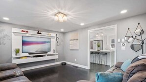 A modern living room with a flat-screen TV, dark flooring, a sofa, and a kitchen area visible through an open doorway. - Sakura House: Spacious 4BR w/ Gym & Premium Design (Northborough)