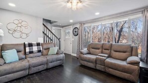 A living room with two sofas, a chandelier, a clock, and a view of trees through large windows. - Sakura House: Spacious 4BR w/ Gym & Premium Design (Northborough)
