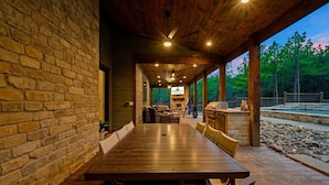A covered outdoor dining area with a long wooden table and chairs, stone walls, and a view of a natural landscape. - Rare Breed | 8 Bedrooms | Pool | Pickleball | Playset | ATV access (Broken Bow)