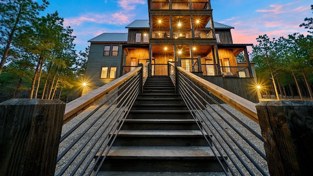 A multi-story wooden house with a staircase leading to the entrance, surrounded by trees. - Rare Breed | 8 Bedrooms | Pool | Pickleball | Playset | ATV access (Broken Bow)
