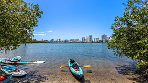 Una orilla tranquila con kayak y tablas de remo, cielo despejado y el perfil distante de una ciudad.