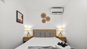 Una cama bien hecha con una colcha gris, una lámpara de noche y un aire acondicionado de pared.