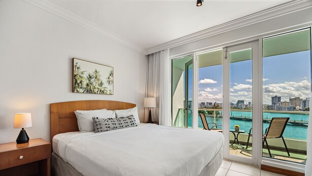 A hotel room with a large bed, a nightstand, a lamp, a painting, and a balcony with a view of the city and water. - Oceanfront Terrace Condado Near The Beach (San Juan)