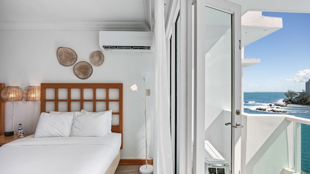 A bedroom with a bed, bedside lamps, a wall-mounted air conditioner, and a view of the ocean through a balcony door. - Condado Beach View Balcony Condado Beach (San Juan)