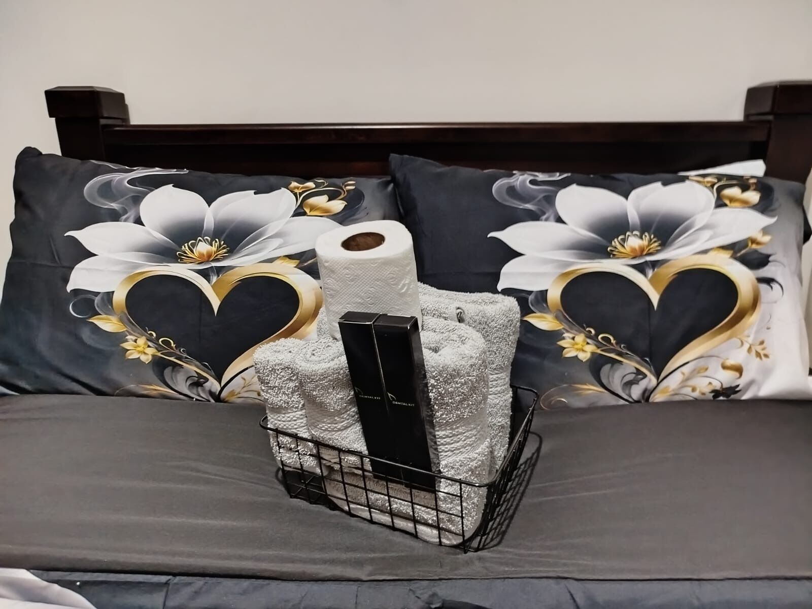 A bed with dark bedding, two pillows with floral and heart designs, a tissue box, and a tissue roll.