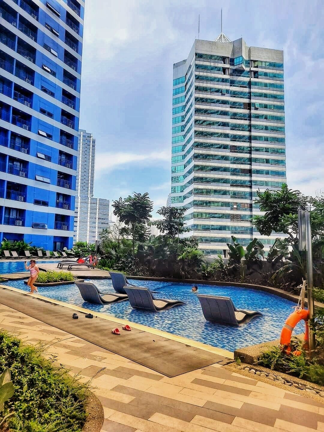 Area piscina con lettini, un galleggiante e alti edifici sullo sfondo.
