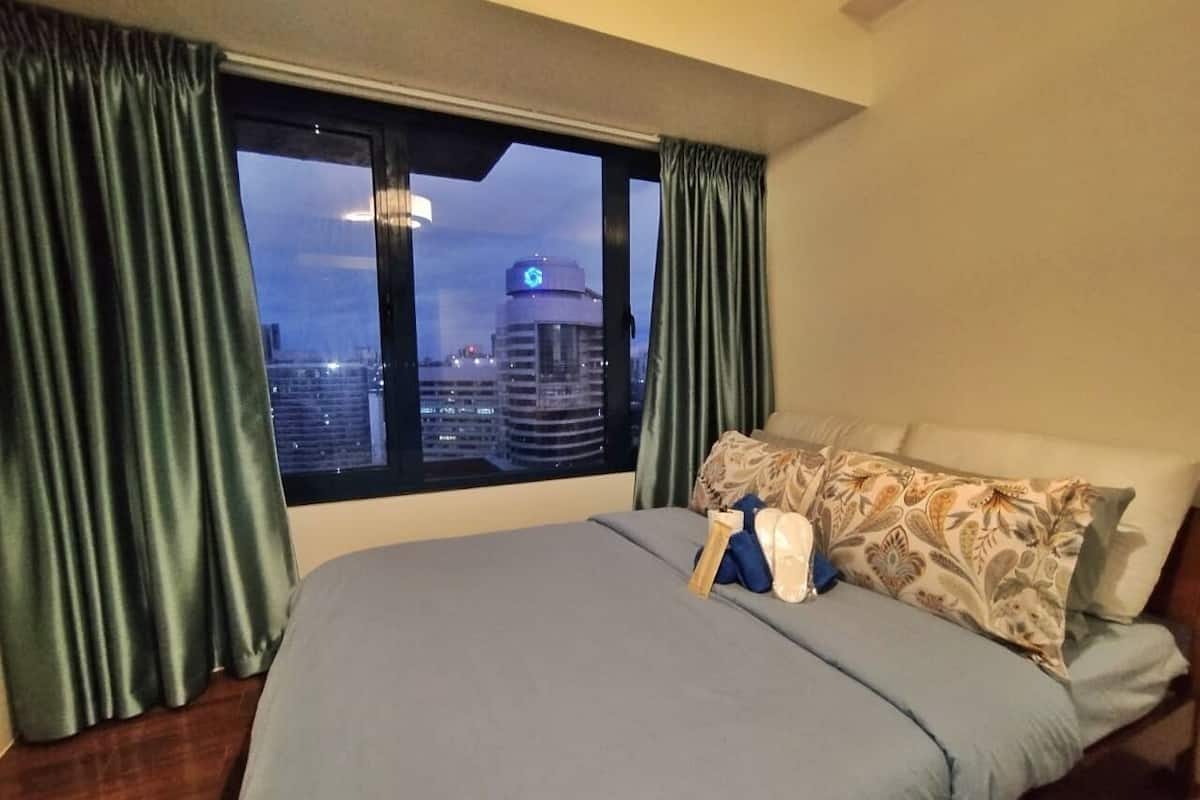 A hotel room with a large bed, green curtains, and a view of a cityscape at dusk.