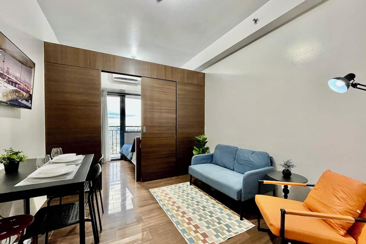 A modern living room with a sofa, a dining table set, a patterned rug, and a balcony with a view.