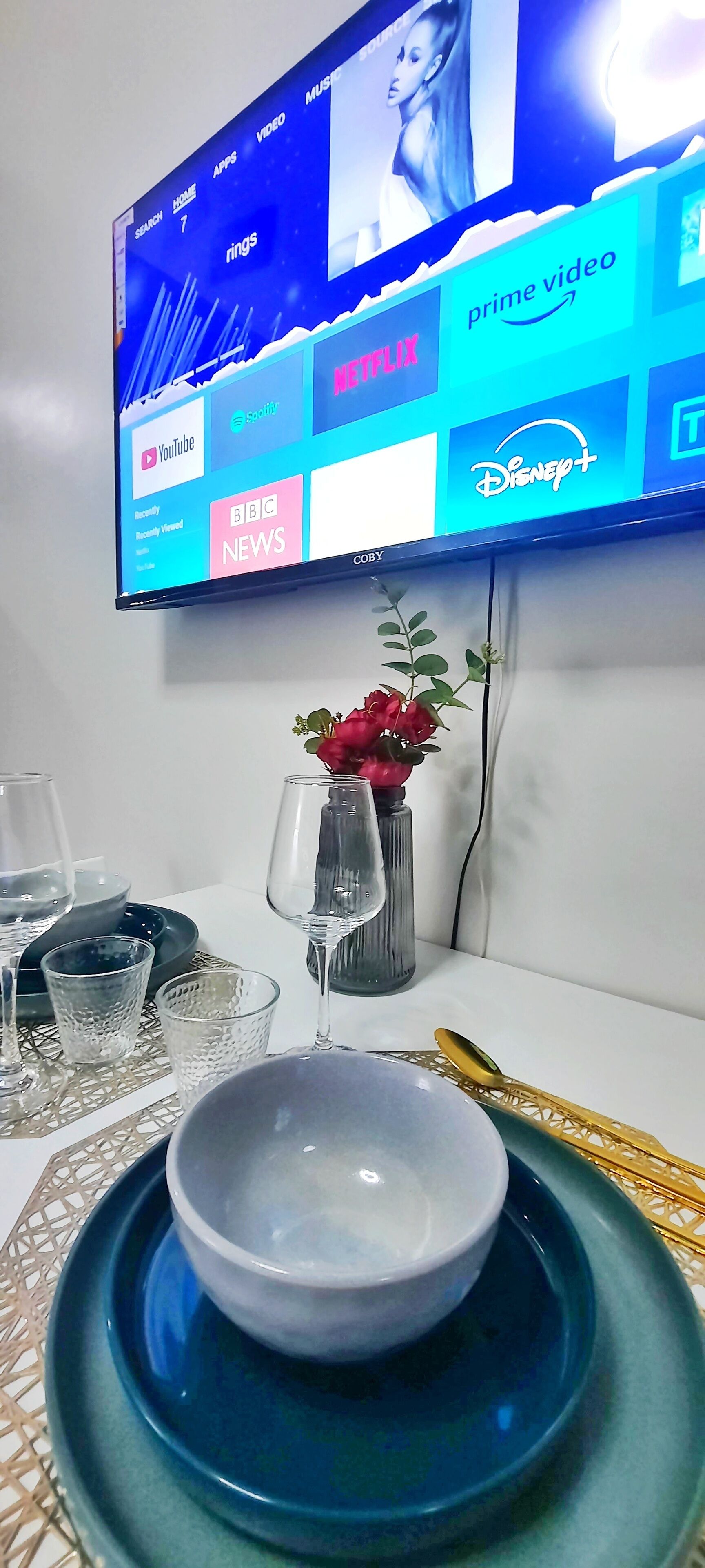 Un comedor con una mesa lista para dos personas, un jarrón con flores rojas y un televisor de pantalla plana mostrando la interfaz de un servicio de streaming.