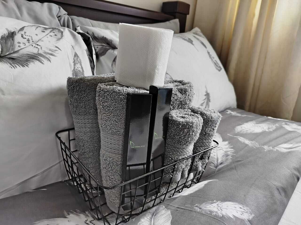 Una cama con una colcha gris estampada, una mesita de noche de metal negro y una bandeja negra con un dispensador de pañuelos blancos y dos toallas negras.