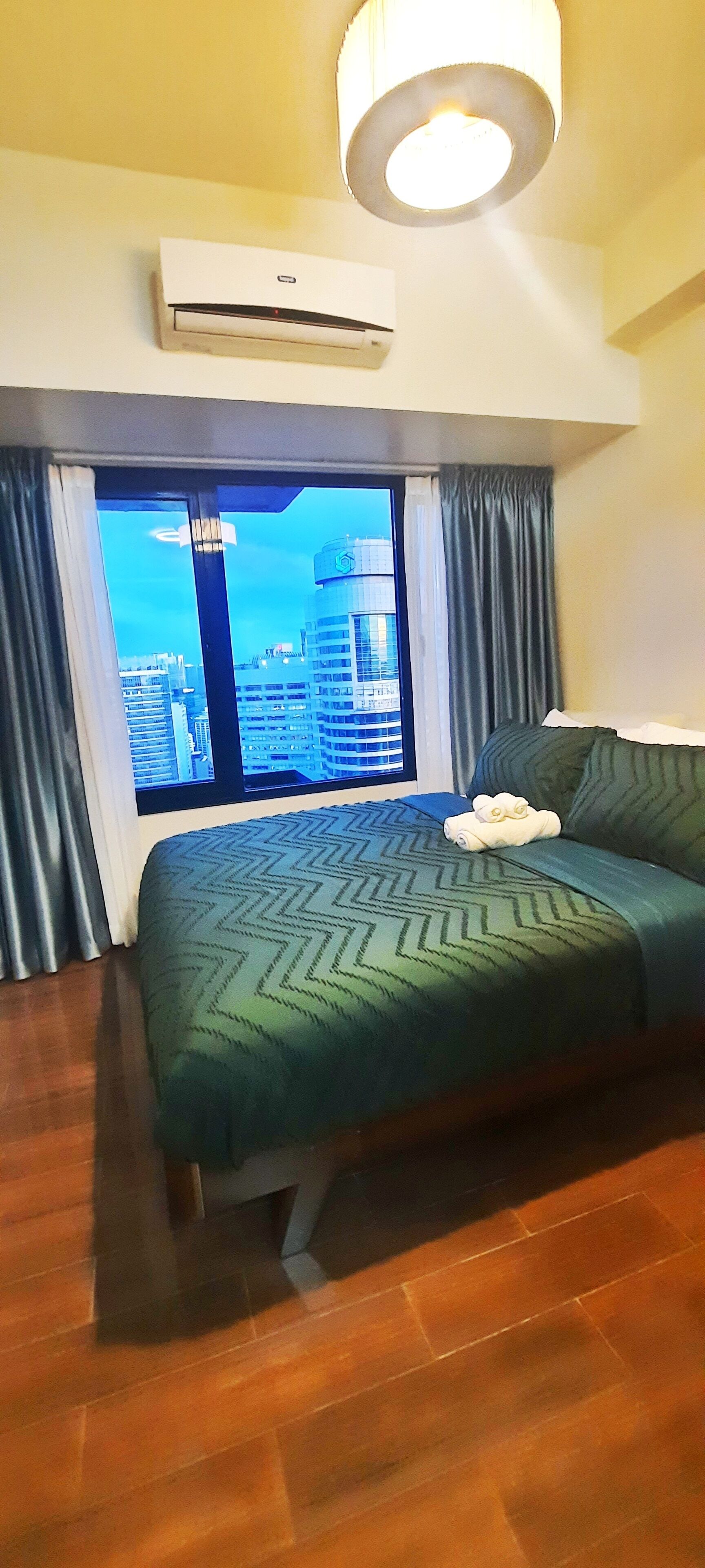 Habitación de hotel con una cama grande, vistas de la ciudad a través de una ventana, un equipo de aire acondicionado y una lámpara en el techo.