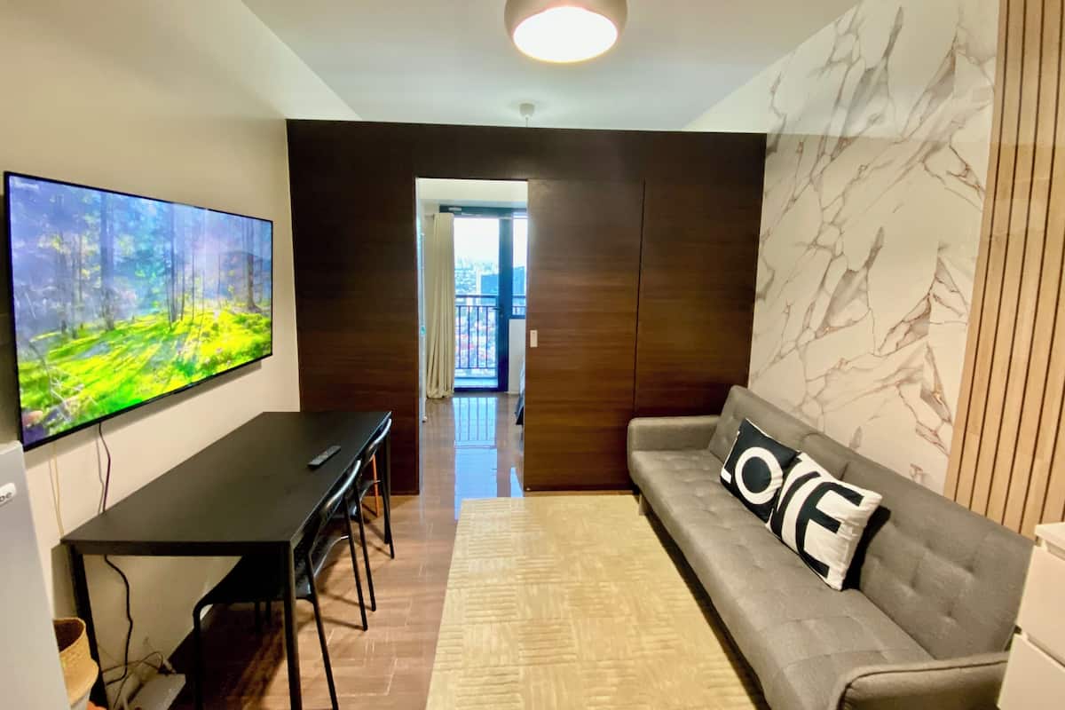 A modern living room with a flat-screen TV, a sofa with a 'LOVE' pillow, a dining table with chairs, and a view into the apartment.