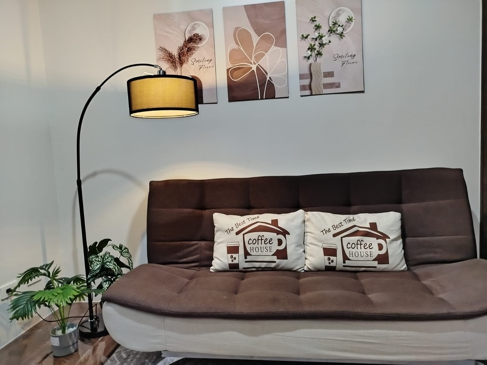 Un sofá marrón con dos almohadones decorativos, una lámpara de pie y un cuadro en la pared.