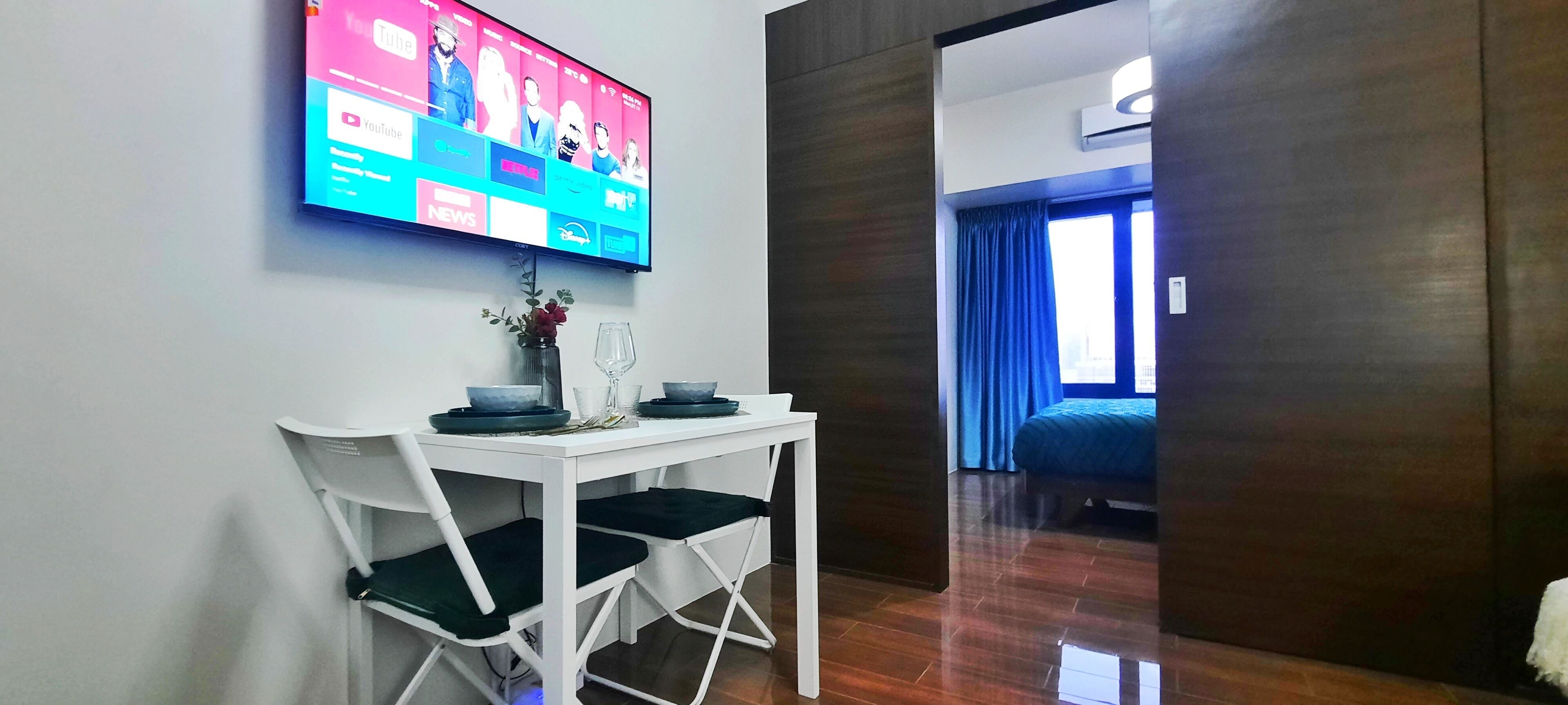 Habitación de hotel con una televisión instalada en la pared mostrando un canal de YouTube, una mesita con un florero y una copa de vino, y vistas de la ciudad a través de una ventana con cortinas azules.