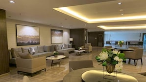 A modern hotel lobby with a sofa set, a round table with a vase of flowers, and a dining area with chairs.