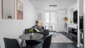 A modern living room with a sofa, a dining table, and a TV. There is a balcony with a view of the city. - Apartments Deluxe Barcelona 55 (Kraków)