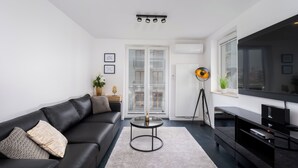 A modern living room with a black leather sofa, a round coffee table, a flat-screen TV, and a large window with a view of buildings. - Apartments Deluxe Barcelona 55 (Kraków)