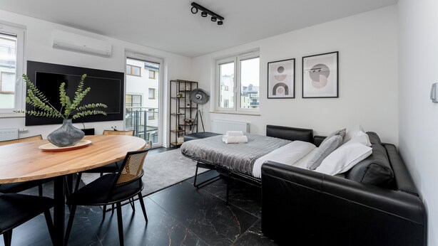 Ein moderner, hell erleuchteter Raum mit Bett, einer Couch, einem Esstisch mit Stühlen, einem Fernseher und Bildern an den Wänden.