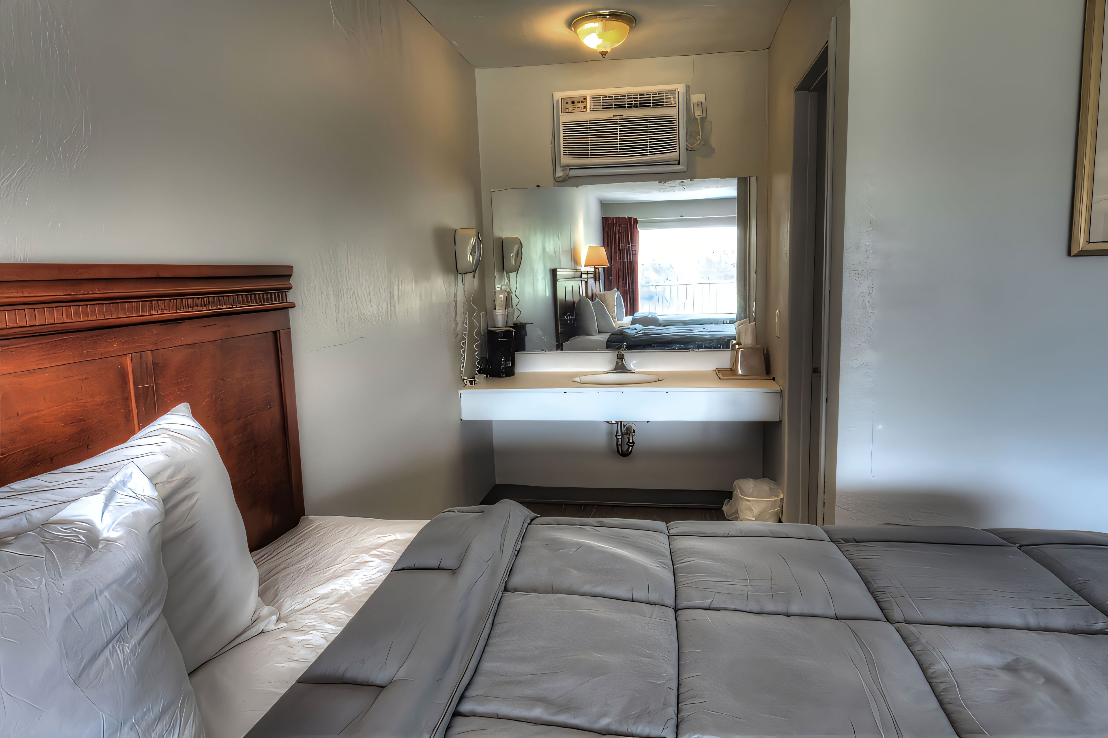 A hotel room with a bed, a wooden headboard, a bathroom with a sink and mirror, and a window with a view.