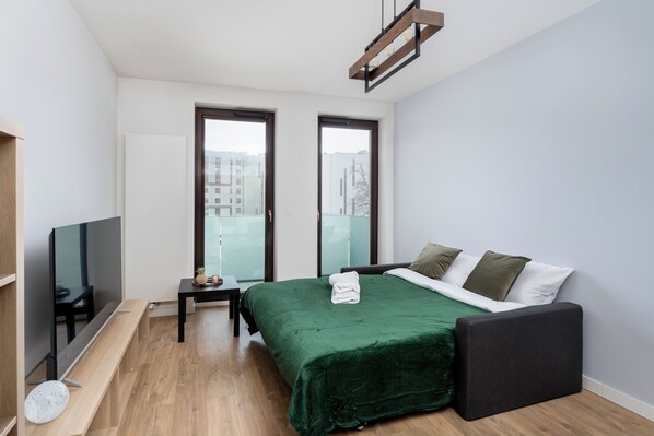 A modern bedroom with a green bedspread, a black bench, a TV on a stand, and large windows with a city view.