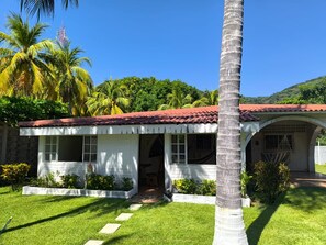 A white building with a red-tiled roof, surrounded by lush greenery and palm trees. - Golden Sand: Beach House Escape, Private Pool, Sea Steps Away (Santa Isabel Ishuatan)