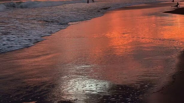 Una playa con un atardecer, olas y algunas personas caminando.