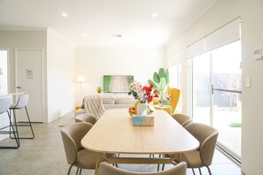 A modern dining area with a long table, multiple chairs, a vase with flowers, and a sofa in the background.