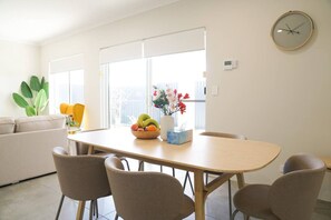 A modern living room with a sofa, a dining table with chairs, a fruit bowl, and a vase with flowers.
