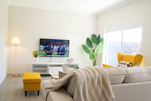 A modern living room with a sofa, a yellow ottoman, a TV mounted on the wall, a floor lamp, and a large green plant.