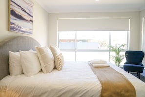 A hotel room with a large bed, a headboard, a window with blinds, a blue chair, and a painting on the wall.
