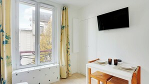 Ein Zimmer mit Fenster, einem Flachbildfernseher an der Wand, einem Esstisch mit Stühlen und einem Heizkörper unter dem Fenster.