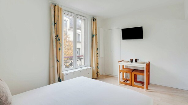Ein Schlafzimmer mit Bett, Schreibtisch mit Stühlen, einem an der Wand montierten Fernseher und einem Fenster mit Vorhängen.