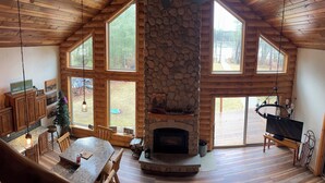 Amplia sala de estar con chimenea de piedra, techo de madera y ventanas grandes que ofrecen vistas al exterior.