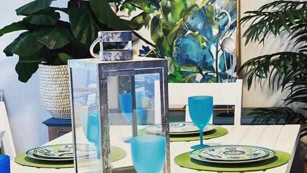 Uma mesa de jantar posta com copos azuis e pratos estampados, rodeada por jogos americanos verdes e um candeeiro decorativo.