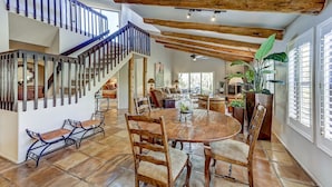 Un vaste espace dédié à la salle à manger, avec une grande table et des chaises en bois, un escalier doté d’une rampe bleue et de grandes fenêtres équipées de volets.