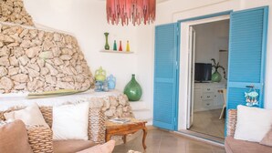 Un saló amb mobles de vimet, una paret de pedra, una porta blava, i una cuina que es veu a través de la porta.