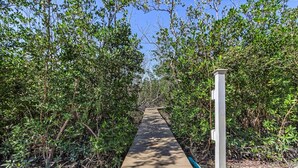 Un sentier en bois bordé d’une végétation luxuriante, qui mène à une construction dotée d’une colonne blanche.