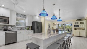 Une cuisine moderne avec des meubles blancs, un grand îlot central doté de tabourets, des suspensions bleues et un réfrigérateur en acier inoxydable.