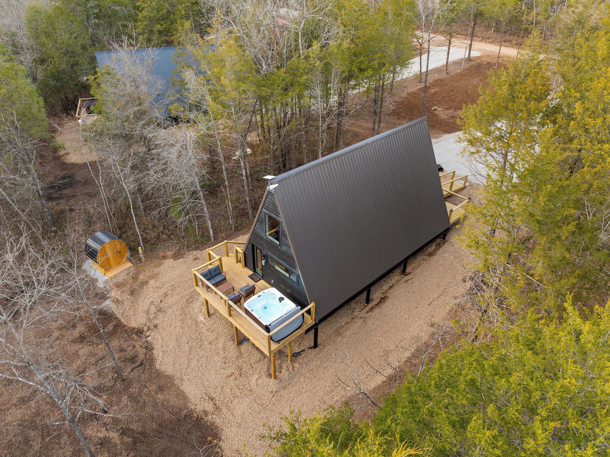 A modern, triangular-shaped cabin with a deck and hot tub, surrounded by trees and a dirt path.