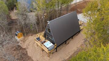 A modern, triangular-shaped cabin with a deck and hot tub, surrounded by trees and a dirt path.