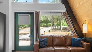 A modern interior with a leather sofa, a wooden sloped ceiling, and a view of a forested area through a glass door. - LEDCabin, See the Stars, w/private Sauna, Hot Tub, on Creekfront (Omaha)