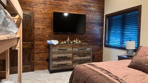 A bedroom with a wooden wall, a flat-screen TV mounted above a dresser, a window with blinds, and a bed with a brown comforter. - Buffalo Creek at Ponds Lodge Cabin 26 (Island Park)