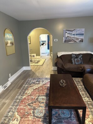 Een woonkamer met een bruine bank, een salontafel en een vloerkleed. Achtergrond: een keuken met uitzicht op het strand.