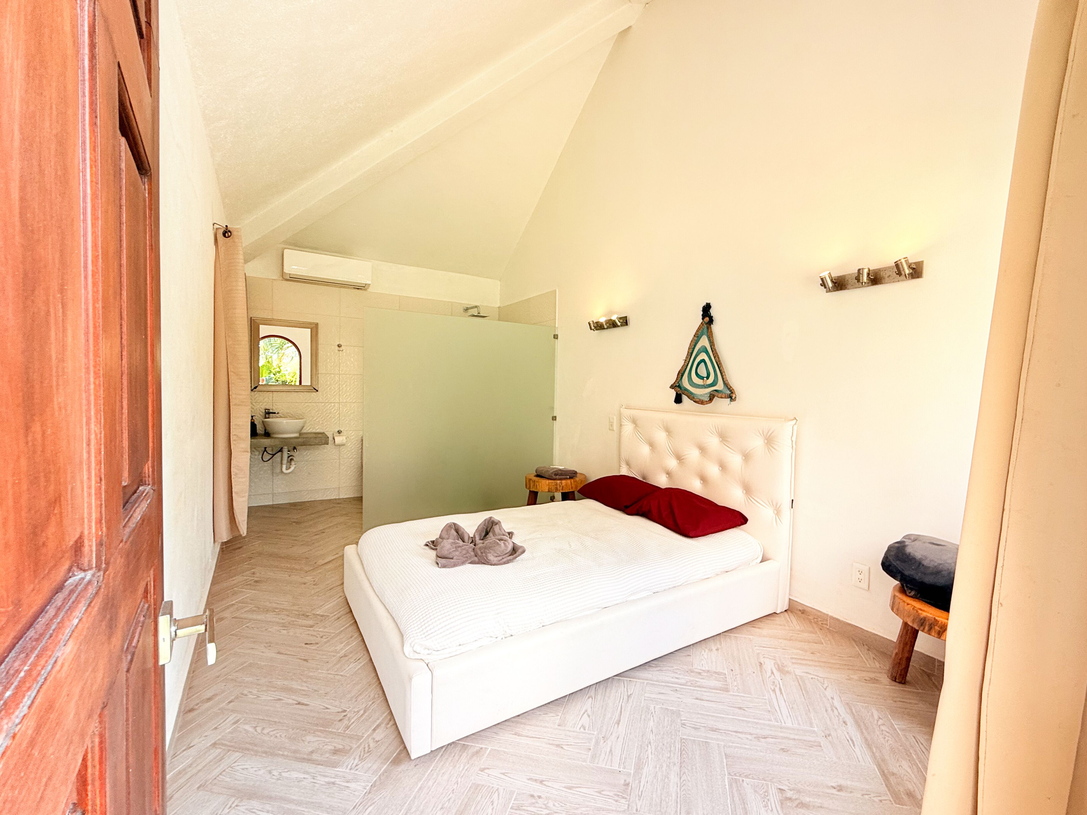 A bedroom with a white bed, a red pillow, a wooden floor, and a bathroom visible through an open door.