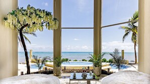 Une chambre avec vue sur la plage, dotée d’une grande fenêtre en arc de cercle, d’une petite table avec une théière et de deux fauteuils blancs moelleux.